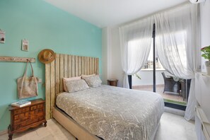 Quarto com cama, cabeceira de madeira, mesa de cabeceira, cômoda, chapéu e vista para o exterior.