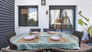 Una mesa redonda con mantel estampado, rodeada de sillas de mimbre, frente a una pared blanca con una ventana y una puerta.
