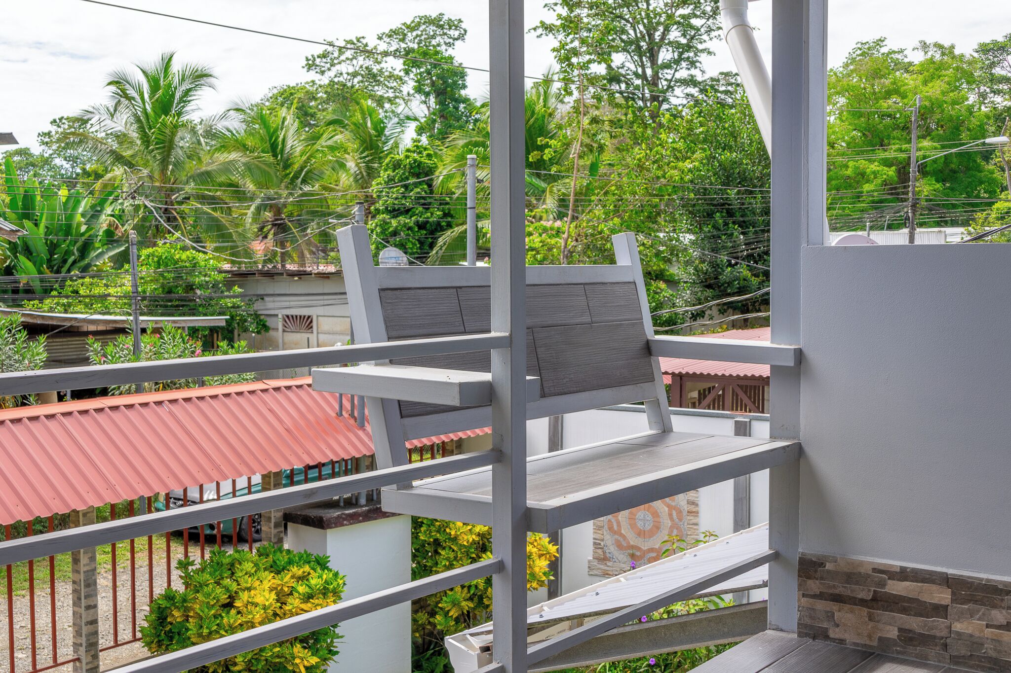 A balcony with a metal railing, a chair, and a view of a residential area with trees and buildings.