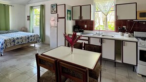 A compact room with a bed, a dining table with chairs, a kitchenette, and a window with curtains. - Villas Cozy Sloth (Puerto Viejo de Talamanca)