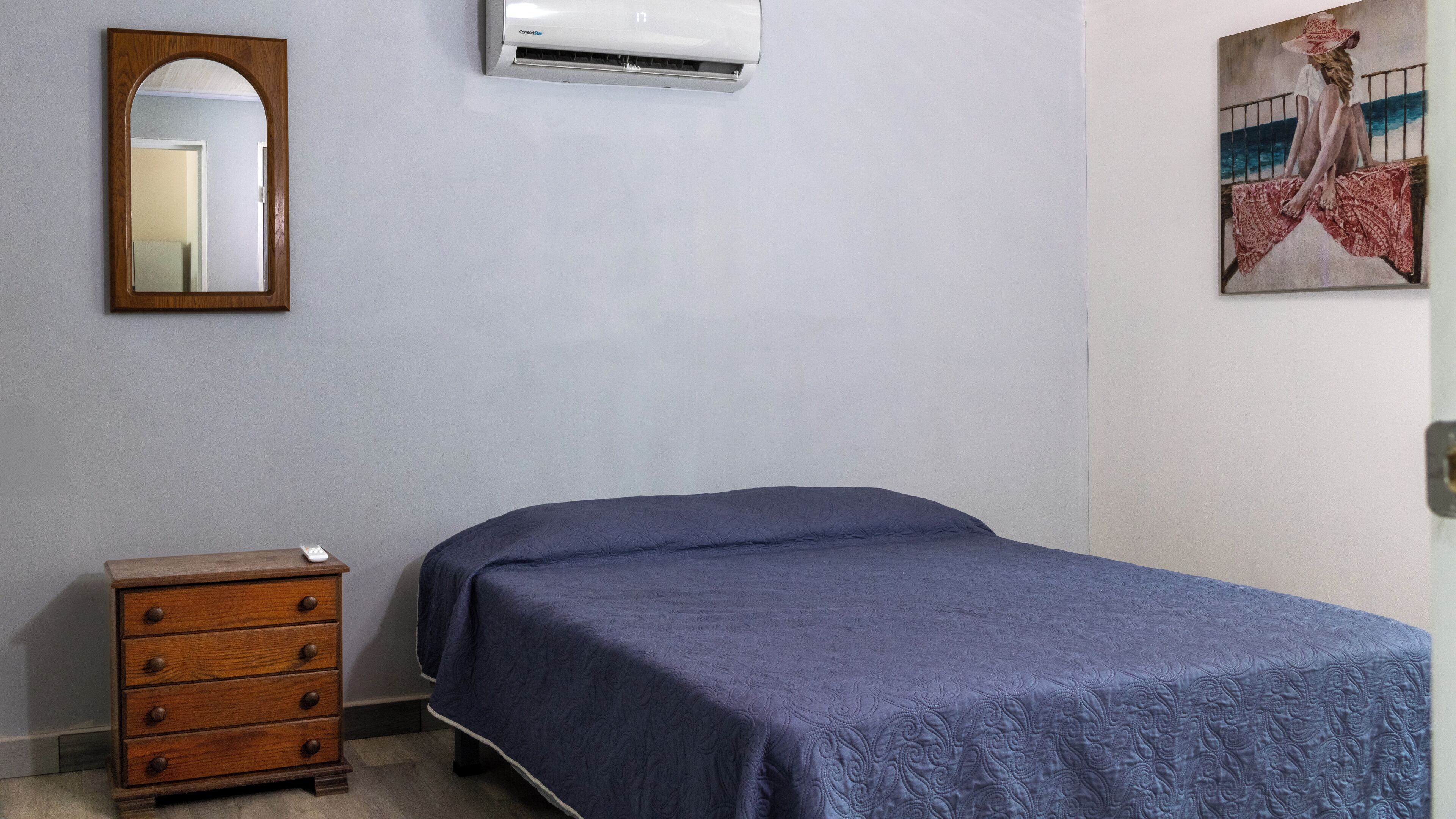 A bedroom with a bed, dresser, air conditioning unit, and a painting on the wall.
