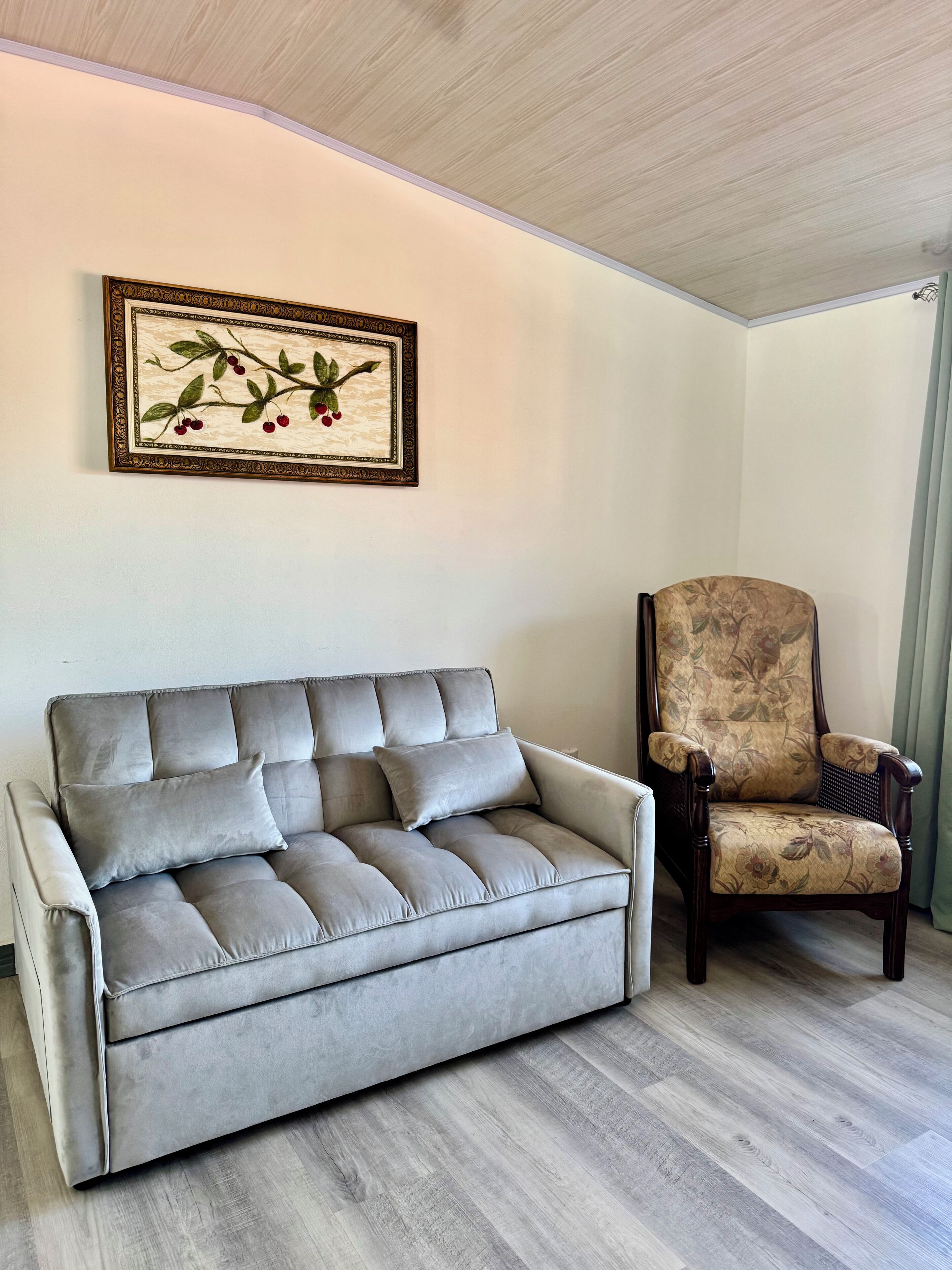 A grey sofa with multiple cushions and a floral-patterned armchair in a room with light wooden flooring and a ceiling with a wood-like texture.