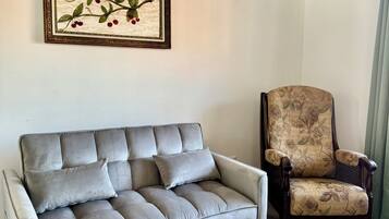 A grey sofa with multiple cushions and a floral-patterned armchair in a room with light wooden flooring and a ceiling with a wood-like texture.