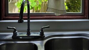 A kitchen sink with a faucet, a window with a curtain, and greenery outside. - Villas Cozy Sloth (Puerto Viejo de Talamanca)