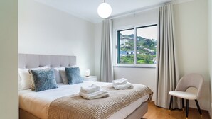 Une chambre d’hôtel moderne avec un grand lit, une vue sur un paysage verdoyant, un fauteuil et une table de chevet avec une lampe.