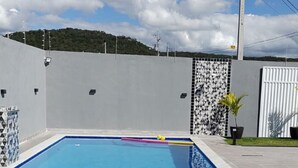 Uma piscina retangular com borda azulejada, escada de metal e um pequeno trecho de parede revestida com padrão geométrico.