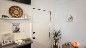 A small, neatly organized room with a bed, a desk, a TV mounted above the door, a small plant, and a heater.