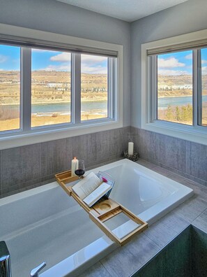 Une salle de bain moderne avec une grande baignoire, deux fenêtres donnant sur une rivière et des collines, ainsi qu’un plateau avec un livre et des bougies.