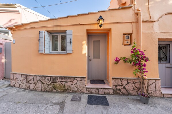 A building with a stone base, a door, a window with shutters, a potted plant, and a wall-mounted light fixture.