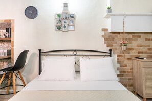 A neatly arranged bedroom with a bed, a chair, a clock, framed pictures, and a brick wall.