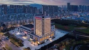 Un moderno edificio de gran altura con varios pisos, rodeado por un estacionamiento y edificios más pequeños.