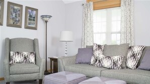 A living room with a grey armchair, a grey sofa with patterned cushions, a floor lamp, and a window with patterned curtains. - Cottage with jacuzzi, Downtown Berlin Ohio (Berlin)