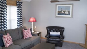 Een woonkamer met een grijze bank, een zwarte leren fauteuil, een rode lamp en een lijst met een afbeelding aan de muur.