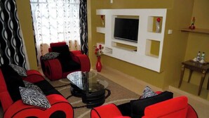 A living room with red sofas, a glass coffee table, a TV mounted on the wall, and a small table with bottles. - Jarika Watamu Breeze Hotel (Nakuru)