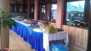 A buffet setup with a long table covered in blue tablecloths, a variety of food items, and a large window with a view of tents outside. - Jarika Watamu Breeze Hotel (Nakuru)
