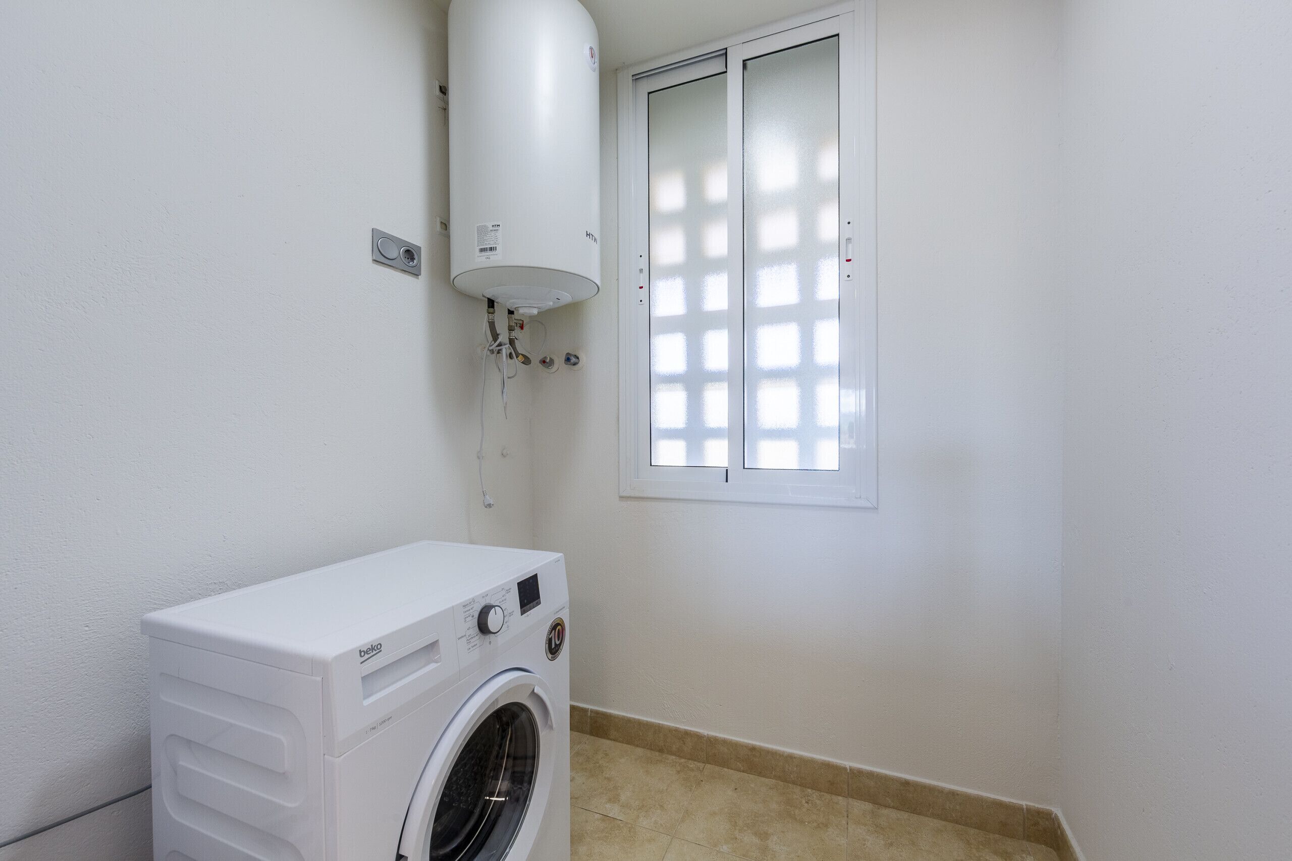 A compact laundry room with a front-loading washing machine, a wall-mounted heater, and a window with blinds.