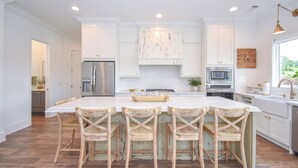 A modern kitchen with white cabinetry, a large island with bar stools, stainless steel appliances, and a view of greenery outside. - Bridge View Retreat, Bridge View Retreat- Luxurious 4Br -Lake Martin Home (Alexander City)
