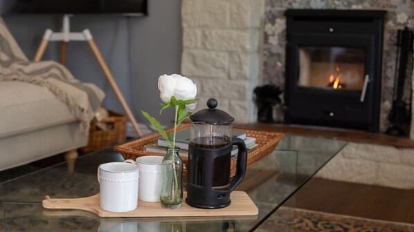 A coffee setup on a glass table with a white flower, a black coffee pot, and two white mugs.