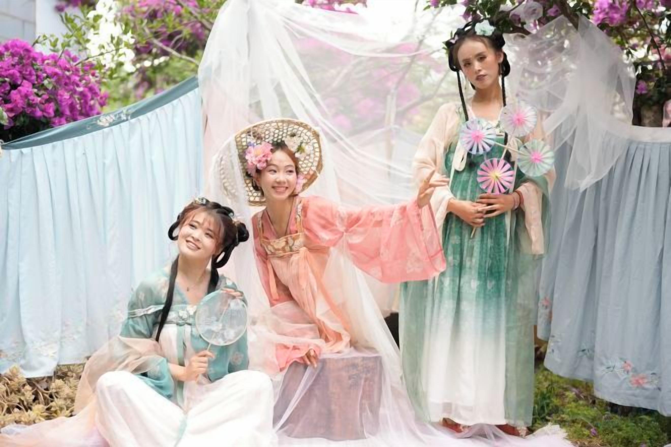 A group of individuals in traditional attire seated and standing in an outdoor setting with a backdrop of draped fabric and floral arrangements.
