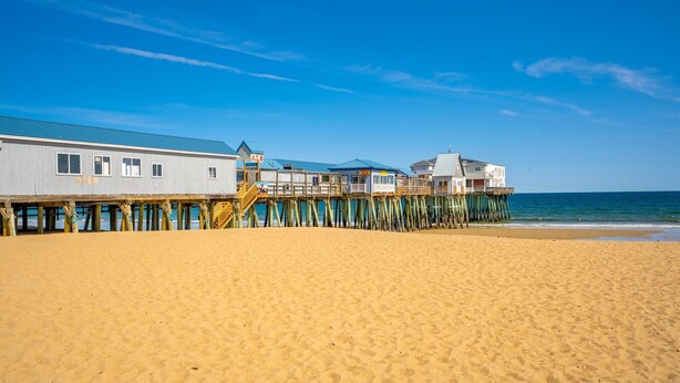 Una playa con un muelle de madera que se adentra en el océano, con varios edificios con balcones y un paseo techado.