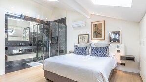 A modern bedroom with a large bed, a glass shower enclosure, a vanity with a mirror, and artwork on the walls.