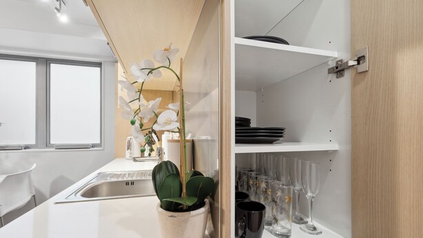 Une cuisine moderne avec un plan de travail blanc, une orchidée en pot et une étagère où sont rangés des verres et des assiettes.