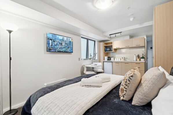A modern hotel room with a bed, a desk, a chair, a kitchenette, and a large window.