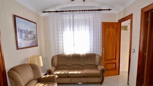 A living room with a sofa, armchair, and a table. There is a window with curtains, a door, and a framed picture on the wall. - Brother's Village (Villardefrades)