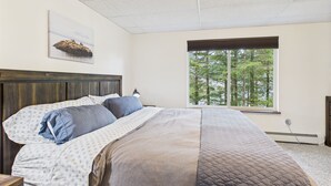 A bedroom with a large bed, a window with a view of trees, a ceiling light, and a wall-mounted picture.