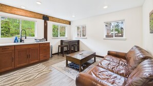 A modern kitchen with a wooden countertop, a sink, and a window overlooking greenery. There is a dining area with a table and chairs, and a leather sofa in the living room.