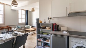 A modern kitchen with a built-in washing machine, a dining area with a table set for two, and a cozy seating area with a sofa and armchair.
