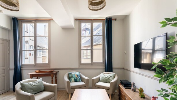 A modern living room with two armchairs, a wooden coffee table, a TV mounted on the wall, and a view of buildings through large windows.