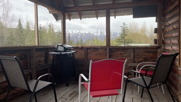 Une cabane en bois dotée d’un porche comprenant deux chaises, un barbecue et offrant une vue sur des montagnes enneigées.