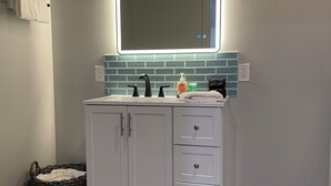 Une salle de bain avec un meuble-lavabo blanc, un miroir avec éclairage indirect, un pare-feu en carrelage et un panier en osier.