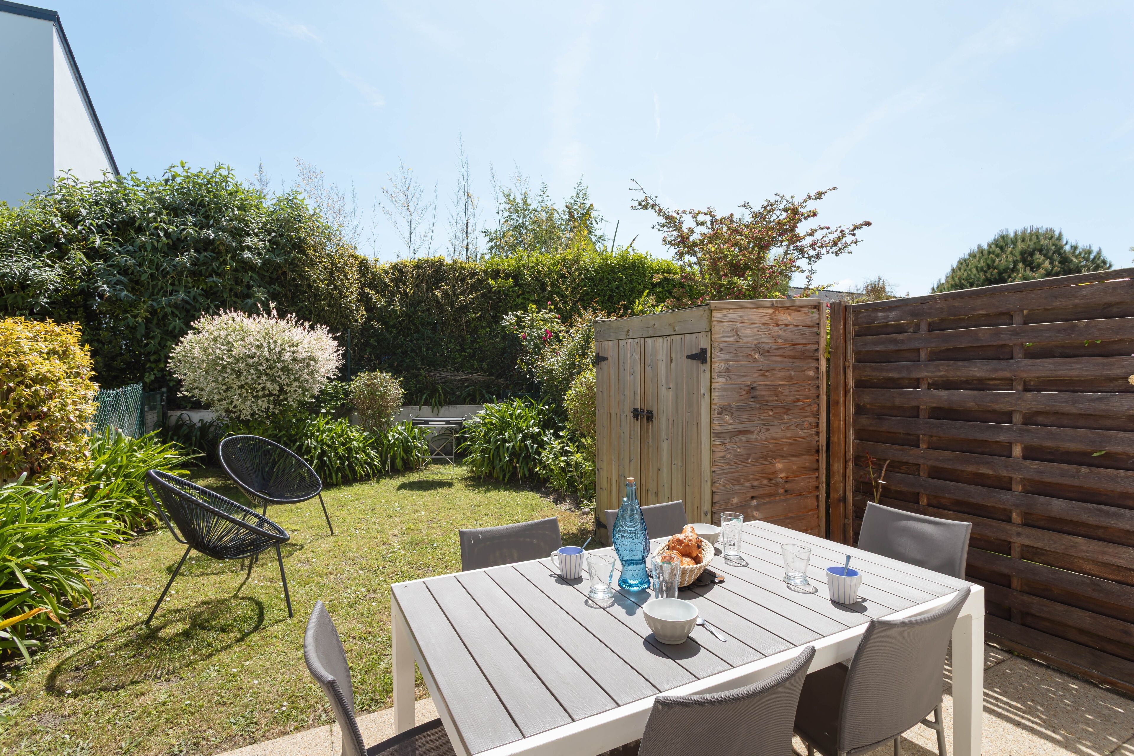 A wooden table with chairs in a backyard setting, surrounded by greenery and a wooden fence.