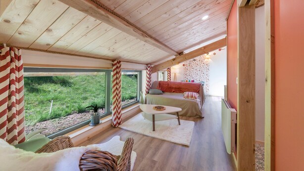A cozy room with a wooden ceiling, a bed with a red blanket, a small table, and a view of a grassy area outside.