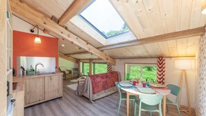 A cozy cabin interior with a wooden ceiling, a skylight, a kitchen area, a dining table, and a bed.
