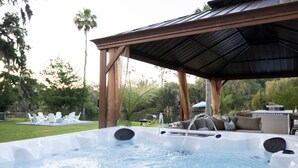 A hot tub with a wooden pergola overhead, surrounded by greenery and outdoor furniture.
