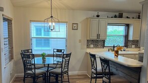 A modern kitchen with a dining area, featuring a wooden floor, a round dining table with chairs, a bar counter, and pendant lighting. - Charming 3-bdrm house w/ community pool + clay tennis courts in Melbourne Beach (Melbourne Beach)