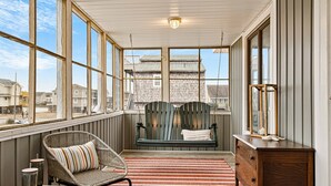 A covered porch with wicker chairs, a wooden bench with cushions, a dresser, and a view of houses through large windows. - Steps to Beach - Playroom - Firepit - Pets OK (Kill Devil Hills)