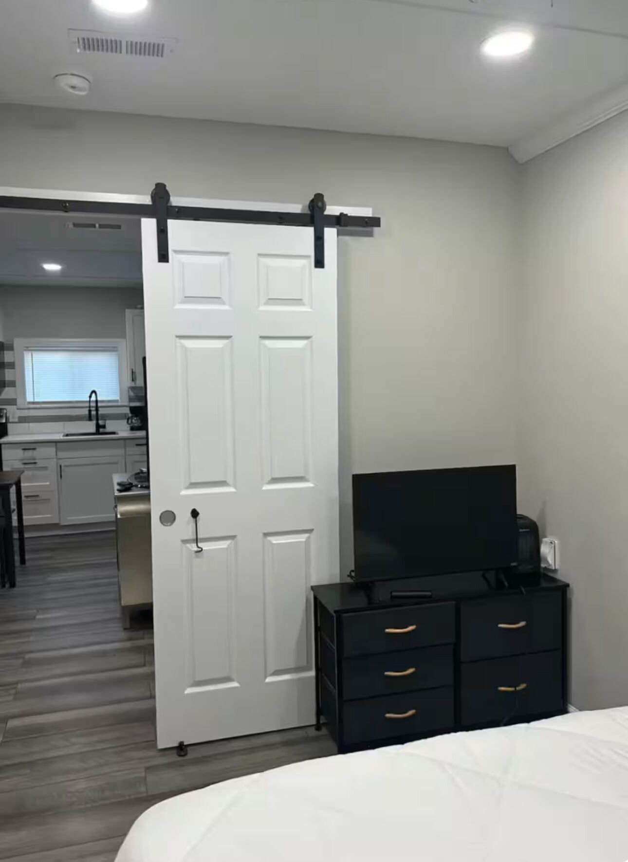 Un dormitorio con cama, televisor, cómoda y puerta corrediza que da a una cocina.