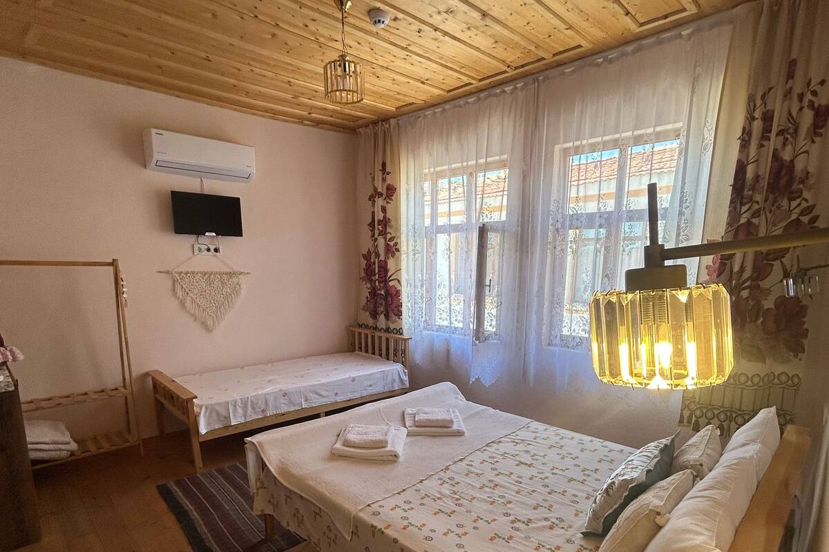 A cozy room with wooden ceilings and walls, a bed with floral bedding, a small bedside table, a TV mounted on the wall, and a window with curtains.