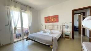 A hotel room with a bed, a bedside table, a fan, a painting on the wall, and a view of the outside through a sliding door.