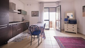 A compact kitchen with dark cabinets, a gas stove, and a dining table with chairs. A large window with curtains leads to an open balcony. - Porticciolo 3 vista mare x 4 persone (Viserba)