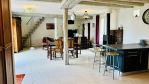 Un salón espacioso con techo alto, vigas de madera, una mesa de comedor con sillas, una zona de cocina con estufa de ladrillo y una escalera que sube a otro nivel.