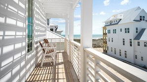 A wooden deck with a white railing, a chair, and a view of a beach and a white building with multiple windows. - Once Upon a Beach 204 COA (Port Aransas)