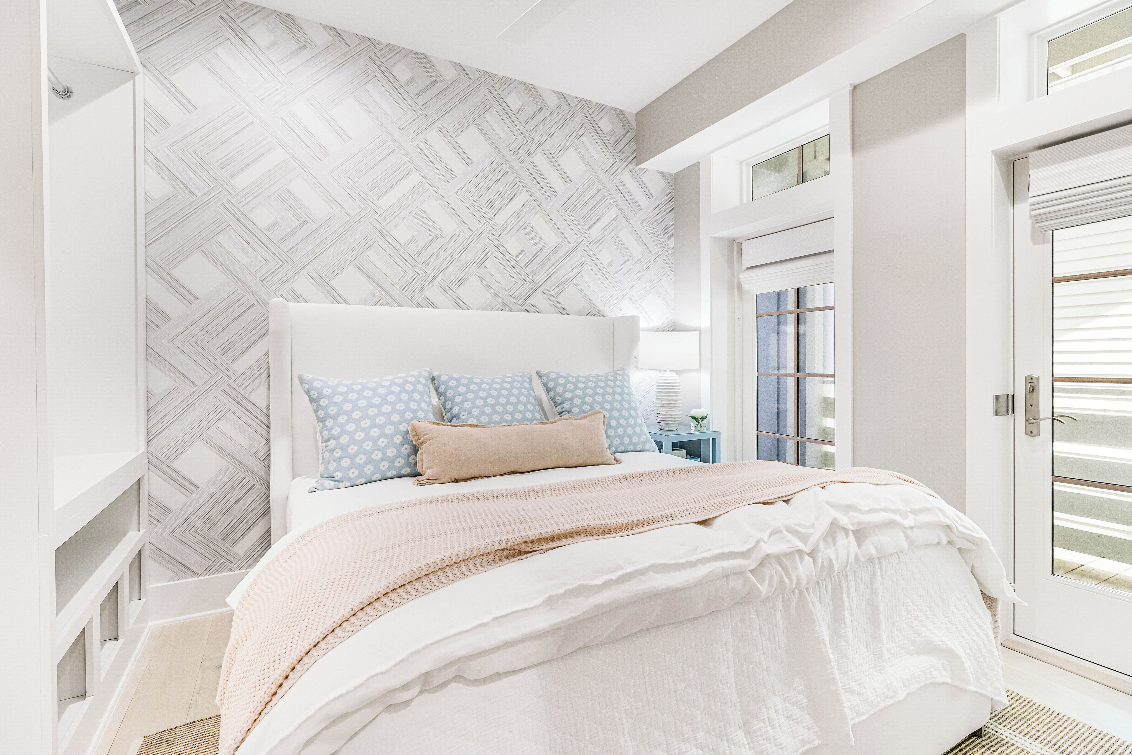 A neatly arranged bedroom with a patterned wall, a bed with a white headboard, and a bedside table with a lamp.