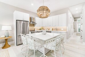 Een moderne keuken met witte kasten, een roestvrijstalen koelkast, een centraal eiland met stoelen en een hangende lamp.