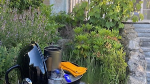 En grill, et gryde og bestik, sat op i en have med en stenvæg og forskellige planter.