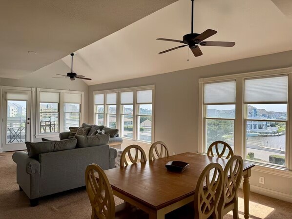 A spacious living room with a gray sofa, wooden dining table, and chairs. There are two ceiling fans, large windows with blinds, and a view of a body of water and buildings outside.